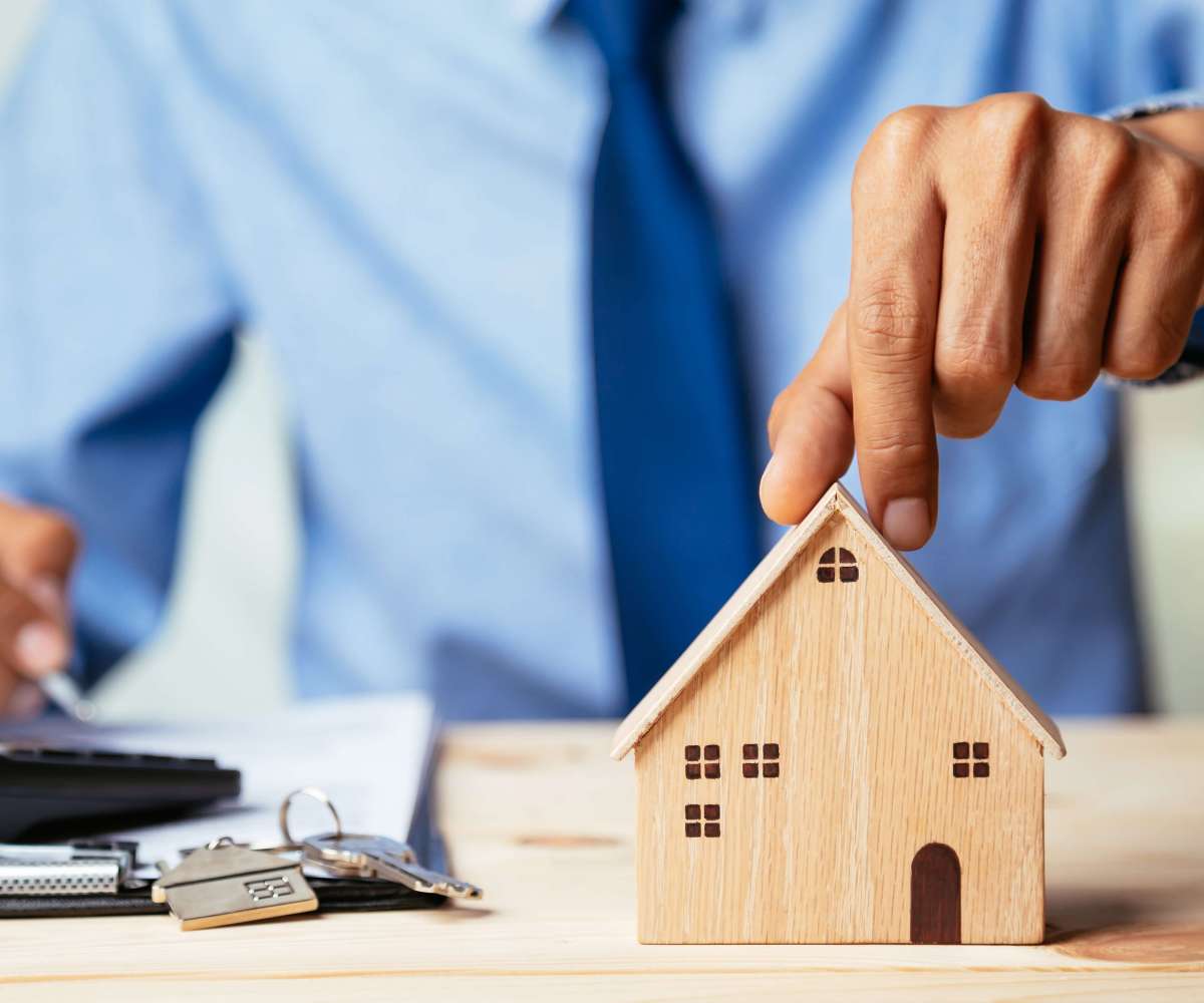 cours immobilier - un agent immobilier pose sa main sur une miniature de maison en bois tout en écrivant sur un document avec une calculatrice et des clés devant lui